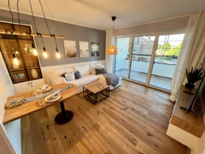 Musterwohnzimmer mit Sofa, kleinem Esstisch, TV-Wand und Zugang zum Balkon